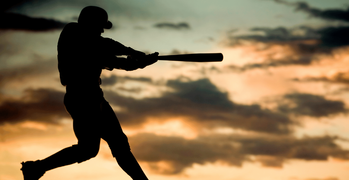 A baseball player swinging his bat behind a cloudy sky near sunset.