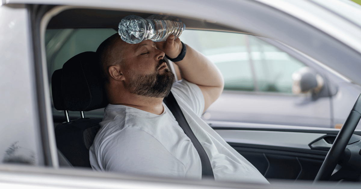 A man sits in the driver's seat of a car, holding a water bottle to his forehead.